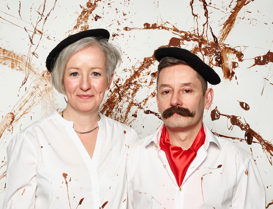 Mark and Emma Baravelli in white shirts and black berets against a chocolate splatter backdrop